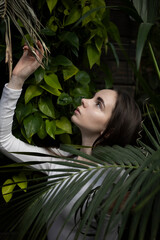 A girl of European appearance, with long dark hair, in a white jacket and jeans, stands in a garden with green leaves. Restricted to Travel, Tropics, Jungle, Pleasure