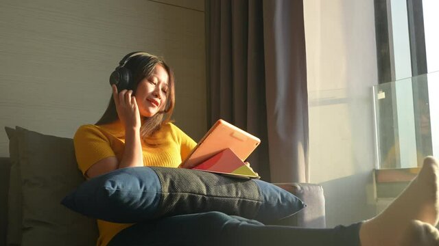 Asian Woman Sit On Sofa Listening To The Music From Headphones While Working On Digital Tablet For Online Business Project At Apartment. Female Freelance Work Job Plan Or Send Online Message At Home.