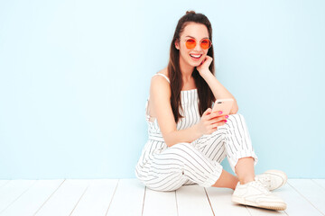 Young beautiful smiling female in trendy summer hipster clothes. Sexy carefree woman posing near light blue wall in studio. Positive model having fun indoors. Enjoying using smartphone apps