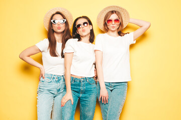 Fototapeta premium Three young beautiful smiling hipster female in trendy summer white t-shirt and jeans clothes.Sexy carefree women posing near yellow wall in studio.Positive models in hats