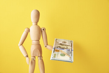 Figure of wooden human holds cash money on yellow background. Wooden doll with dollar banknotes on yellow background. Business and finance concept