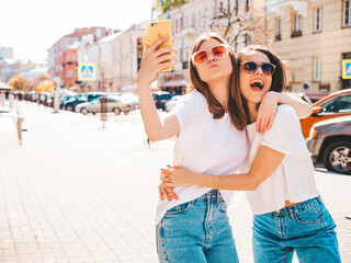 Two young beautiful smiling hipster female in trendy summer white t-shirt clothes and jeans.Sexy carefree women posing on the street background.Positive models having fun, hugging.Taking photo selfie