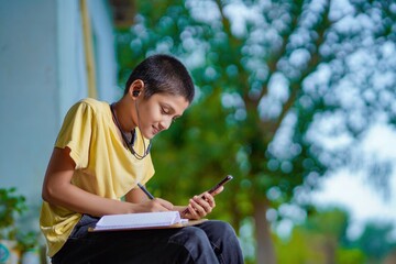 Indian school boy holding phone distance learning class using mobile application, watching online lesson, video calling in app making notes studying at home