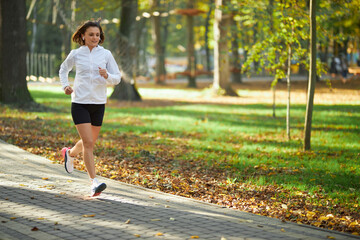 Active young woman in sportswear running at green park during morning time. Happy female with dark...
