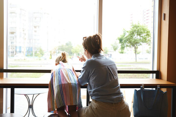 Mother with her little daughter sitting near window in to the cafe. Waiting for the order