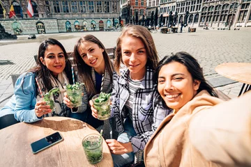 Crédence de cuisine en verre imprimé Anvers temps de selfie. Heureux quatre copines souriantes assises en plein air avec des cocktails en prenant une photo de groupe. mise au point sélective sur le visage de la femme indienne de gauche.