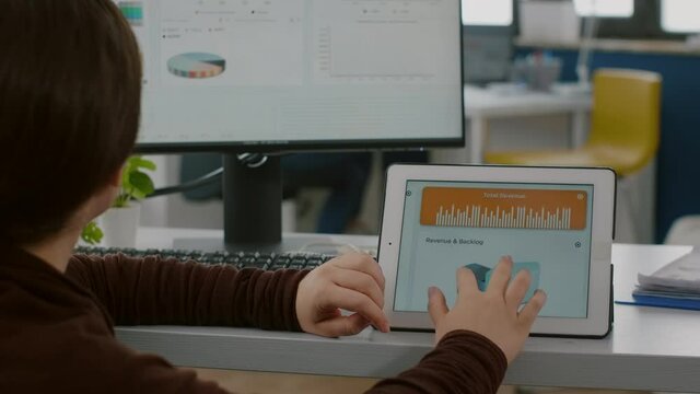 Invalid paralysed handicapped woman entrepreneur looking at computer and tablet checking statistic datas working in business start-up office in wheelchair. Busy employee analysing financial graphs