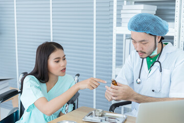 Obraz premium Smart Asian male doctor with white coat is looking at medical tablet pills while diagnosing and talking to beautiful female patient is sitting on wheelchair fort treatment in hospitsl room. 