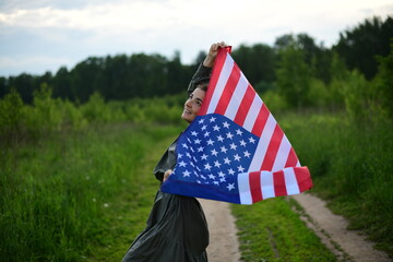 A young, happy American woman with long hair, holding the US national flag fluttering in the wind on her shoulders, is relaxing in the open air, enjoying a warm summer day. July 4
