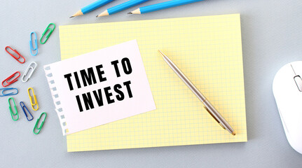 TIME TO INVEST is written on a piece of paper that lies on a notebook next to office supplies.
