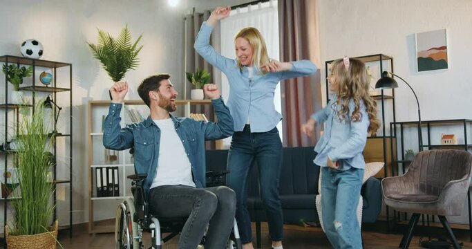 Good-looking smiling happy bearded disabled man in wheelchair watching how dancing his cheerful lovely wife and cute 10-aged daughter ,family leisure concept