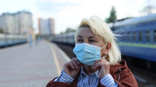 Adult blonde woman tourist puts on protective medical mask on her face on public train station platform during coronavirus Covid-19 pandemic. Travel by train