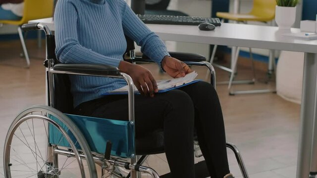 Black woman with disabilities, invalid, disabled paralized handicapped sitting immobilized in wheelchair holding clipboard with financial documents. African manager working in business office room