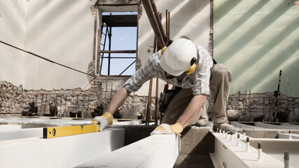 man at work, construction worker wears helmet and uses the spirit level, check the beams at the base of the foundations of the second floor of house, in renovation building site background