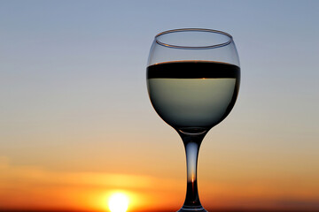 Glass with white wine on beautiful sunset background, sun and sky are reflected in alcohol drink. Concept of celebration, evening party at resort, romantic dinner outdoors