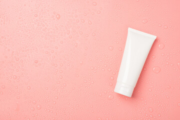 Top view photo of white cream tube without label on the right and water drops on isolated light pink background with copyspace on the left