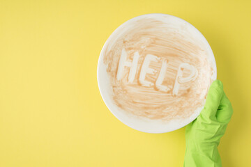 Top view photo of hand in green rubber glove holding dirty dish with inscription help on isolated yellow background with copyspace