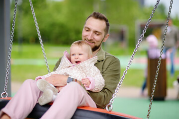 Toddler girl having fun on outdoor playground. Young father rides daughter on swing. Spring/summer/autumn active leisure for family with kids.