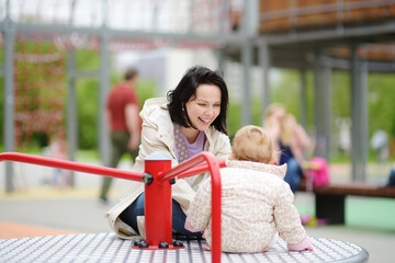 Toddler girl having fun on outdoor playground. Happy young mother rides daughter on carousel. Spring/summer/autumn active leisure for family with kids.