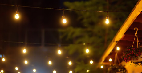 Photo of string lights hanging in restaurant or cafe in the garden at evening time. Fashion decoration with bulbs for night summer party. Outdoor electric lamps