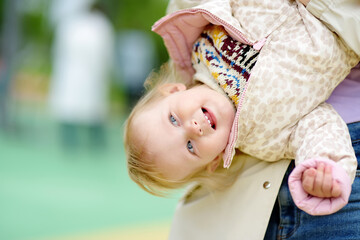 Toddler girl having fun on outdoor playground. Merry young mother swinging her little daughter....