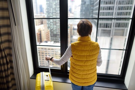 Woman tourist stays in hotel room in New York. Traveler with suitcase looks and admires of view the skyscrapers of Manhattan outside the window. Tourism and travel in USA. Booking apartment