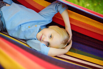 Fototapeta na wymiar Cute little blond caucasian boy having fun with multicolored hammock in backyard or outdoor playground. Summer outdoors active leisure for kids. Child relaxing in hammock.