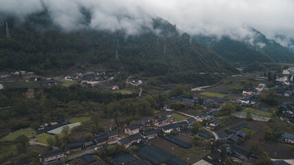 Aerial view of nice Chinese village surrounded by tall mountains. Foggy and cloudy day. Detailed roads and buildings. Town under the low clouds.