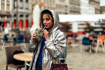 Fototapeta premium girl drinking mojito with straw in city center. young woman in raincoat having a drink in happy hour.