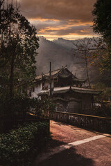On way to Qingcheng mountain, Sichuan, China. Breathtaking golden hour sunset over mountains and old Asian Chinese style building house near road path. Traditional architecture and spears on roof edge