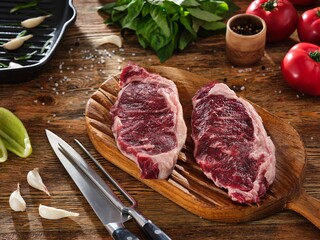 Two juicy raw meat steaks on a wooden cutting board. Knife, tomatoes and spices on the background