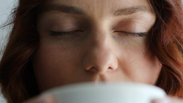 Close up of joyful charming caucasian woman taking a sip of coffee or tea from drink mug in the morning