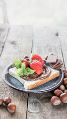 A piece of bread with chocolate paste and nuts paste , heazelnuts and chocolate on a wooden background . 
