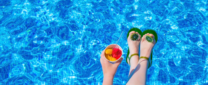 A juicy, summery banner. Women's feet in lime-shaped sandals and a hand with cold ice cream in the pool. Sunlight dancing in the water. The concept of rest, vacation and hot season.