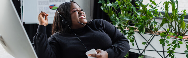 happy african american plus size operator in headset with microphone talking and holding smartphone...