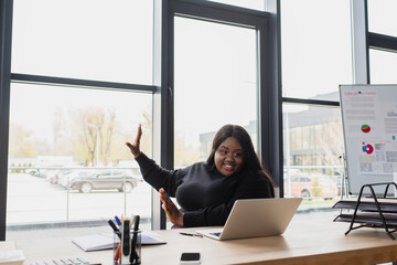 happy african american plus size woman gesturing while having video call on laptop in office.