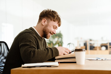 Young ginger man smiling and using mobile phone while reading book