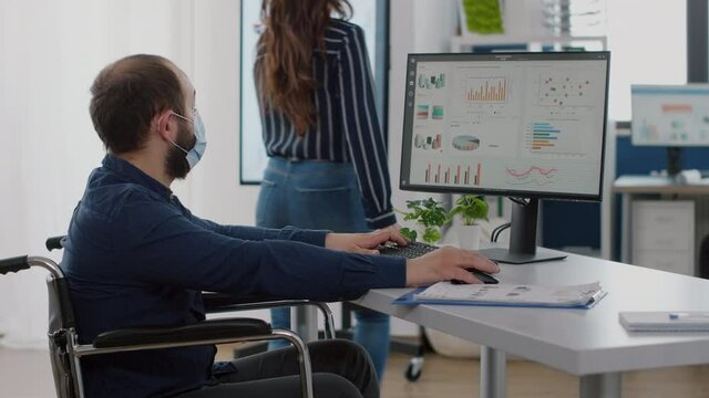 New disabled paralysed invalid employee company staff member with face mask look at computer asking about corporate app program software, manager helping his working in new normal business office