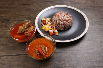 Nasi dagang special brown white rice cooked with coconut milk pickled fruit vegetable pineapple carrot on black offwhite rim plate chicken fish curry in bowl over rustic dark wooden background