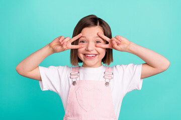 Photo of happy cheerful little girl good mood hold hands fingers face v-sings isolated on teal color background