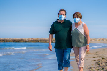 sweet loving mature couple in face mask during covid19 - senior retired husband and wife on their 70s enjoying beach walk relaxed and happy during covid outbreak