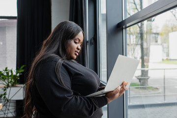 african american plus size businesswoman holding laptop near window in office.