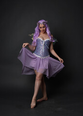 Full length portrait of a purple haired  girl wearing fantasy corset dress with fairy wings and flower crown.  Standing pose against a dark studio background.