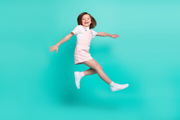 Fototapeta na wymiar Photo of impressed sweet schoolgirl dressed pink overall jumping high smiling isolated turquoise color background