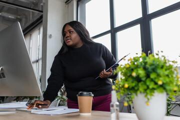 african american plus size businesswoman holding clipboard and looking at computer monitor in office.