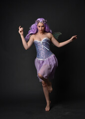 Full length portrait of a purple haired  girl wearing fantasy corset dress with fairy wings and flower crown.  Standing pose against a dark studio background.