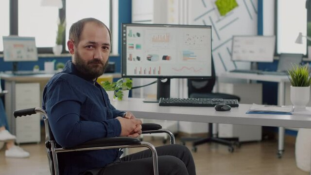 Sad businessman frustrated of his disabilities looking at camera sitting immobilized paralysed in wheelchair in business office working on pc with data, prosessing financial information