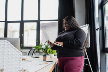 happy african american plus size engineer holding laptop near blueprint and carton house model.