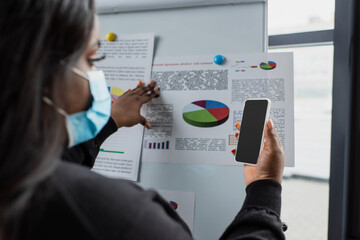 blurred african american plus size businesswoman in medical mask using smartphone near flip chart with charts and graphs.