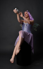 Full length portrait of a purple haired  girl wearing fantasy corset dress with fairy wings and flower crown.  Seated pose against a dark studio background.